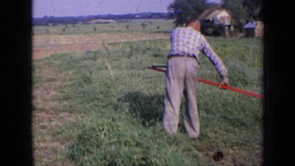 1963 Russia rural field frame showing a man from behind moving a long red-handled tool through low grass near distant farm structures.