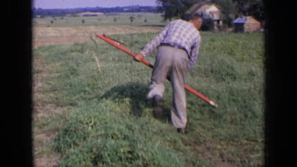 The farmer moves forward through the grass with the long red rake extended behind him in the 1963 rural Russia home-movie frame.