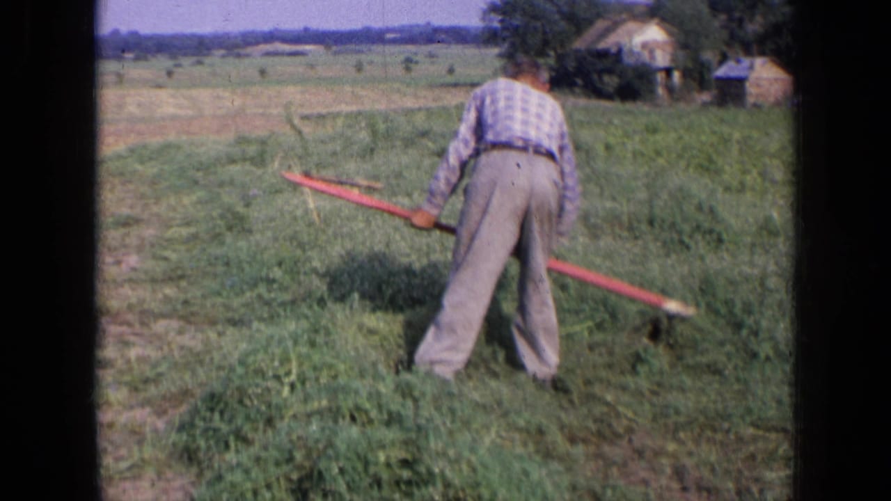 The man continues hand mowing in a grassy field, with low rural buildings visible in the distance.