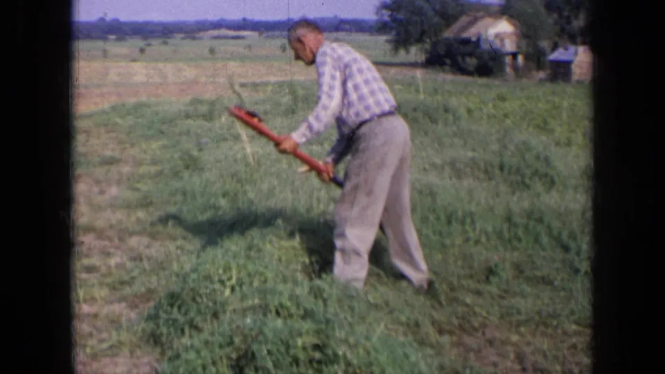 A closer 8mm frame shows the man bending into his work as he cuts hay by hand in the field.