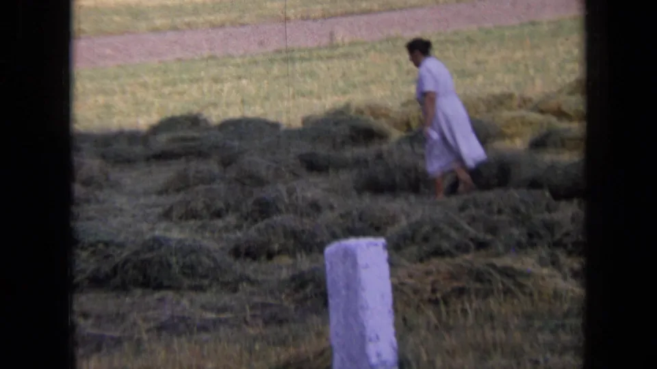 1963 Russia home-movie frame showing a woman in a pale dress moving through cut hay piles in a rural field.