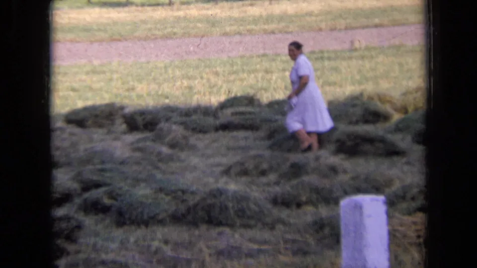 The woman stands among loose hay in a grassy field, with a white post in the foreground and 8mm home-movie borders visible.