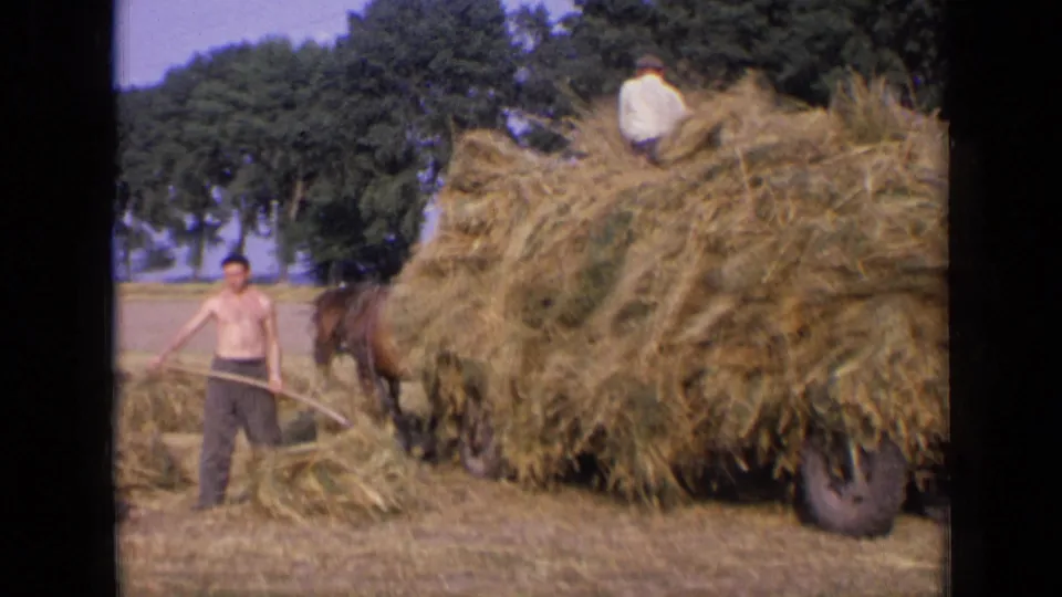 1963 Russia home-movie frame of a shirtless worker holding a pitchfork before an overloaded horse-drawn hay cart.
