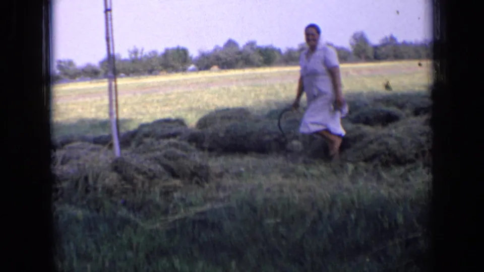 In 1963 Russia home-movie footage, a woman in a white dress stands near piles of cut grass with a hand tool in an open field.
