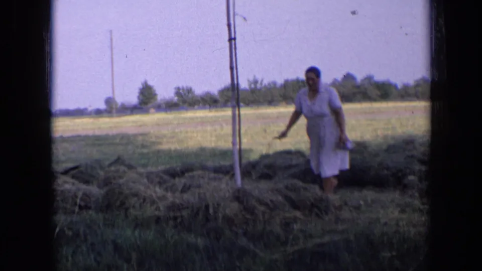 The woman moves through the cut grass piles in an open rural field, framed by trees, poles, and the soft grain of 8mm film.
