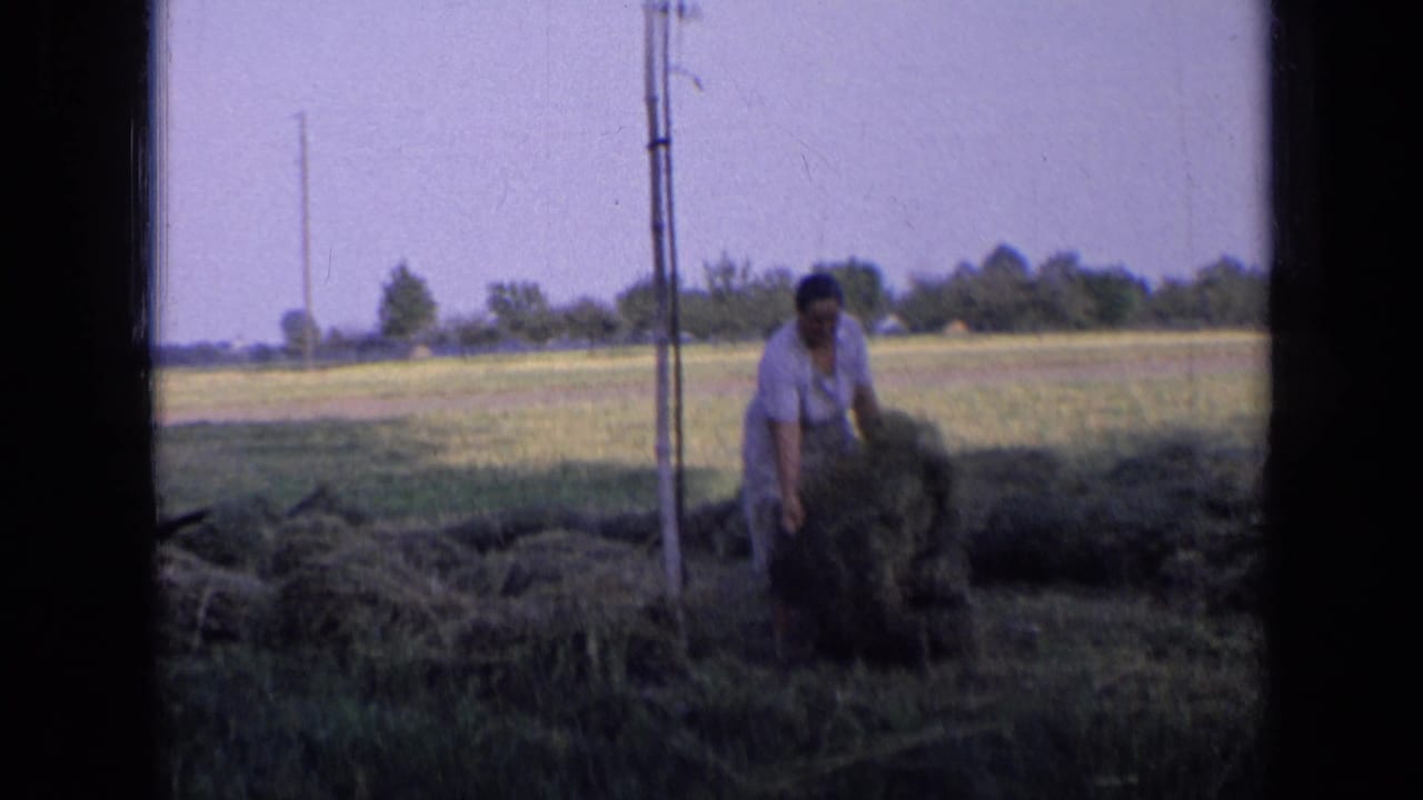 Woman bends near a hay pile with open fields and poles behind her.