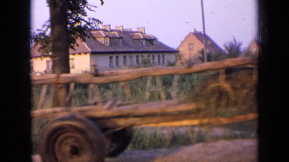 The wooden wagon continues past the camera, showing its side rails, rear wheel, grass, fence, and village buildings beyond.