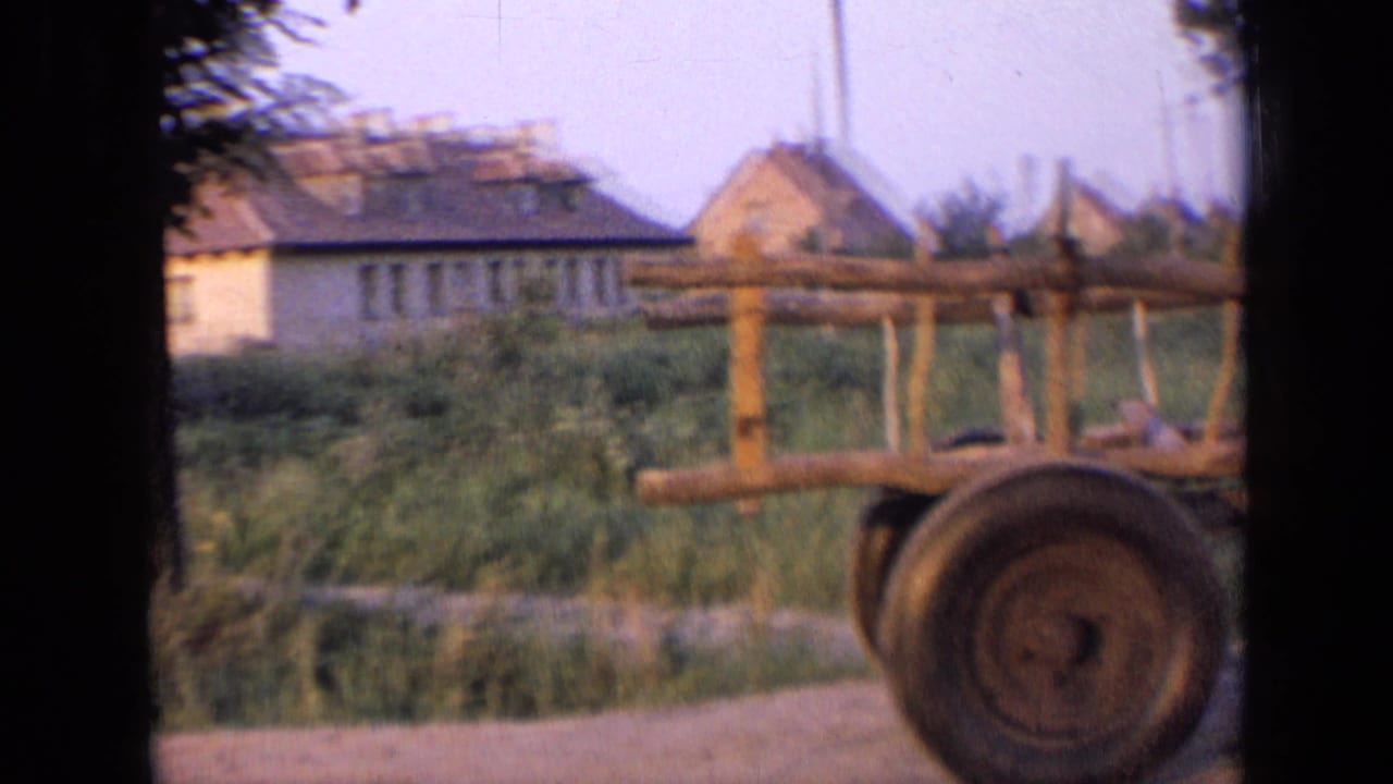 Rural road beside a wooden cart or fence, with houses and greenery in the background.
