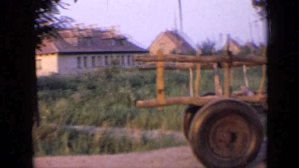 1963 Russia home-movie frame showing a rural roadside, wooden cart or fence, green growth, and distant houses.