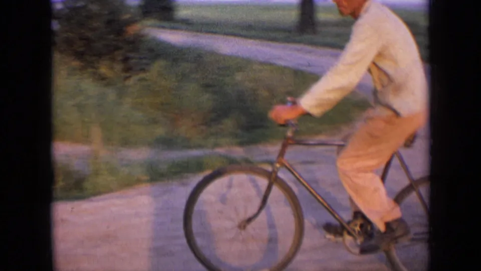 Side view of the cyclist in 1963 Russia, filmed with motion blur as he pedals past grass and a curving rural road.
