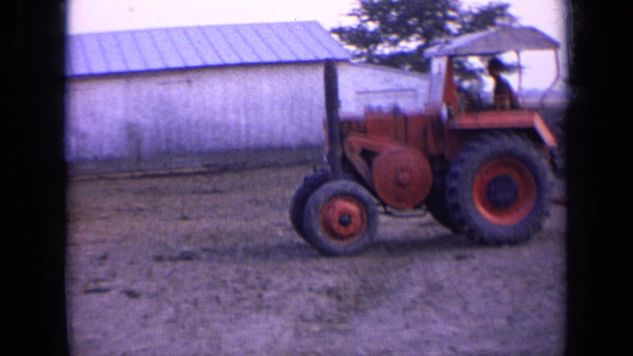 Red tractor driven across a farmyard in front of a pale barn with a metal roof, consistent with the 1963 Russia metadata.