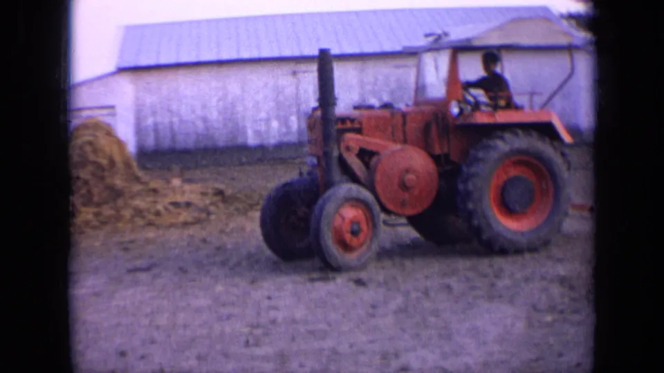 Side view of the red tractor with a seated operator, hay pile, and barn in a rural 1963 Russia farmyard scene.