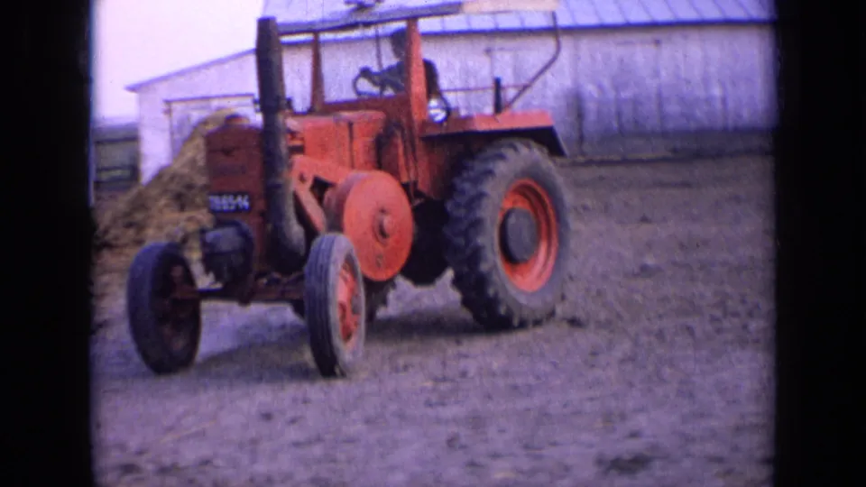 Front angled view of a red tractor with visible markings, a driver, hay pile, and barn in the rural farmyard.