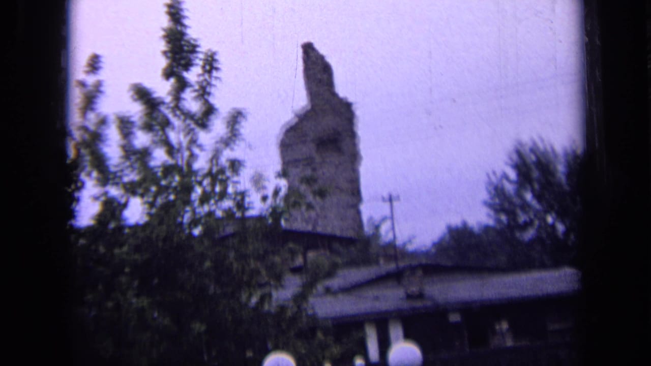 Broken stone tower or stack rising behind trees and a low roof.