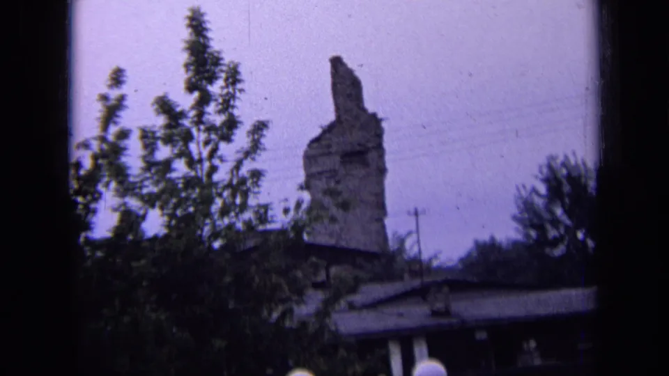1963 Russia 8mm view of a tall damaged stone ruin seen across trees, with a utility pole, roofline, and muted sky.