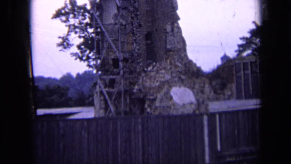 1963 Russia home-movie frame of a large crumbling stone wall or tower base behind a wooden fence, with trees and distant hillside.