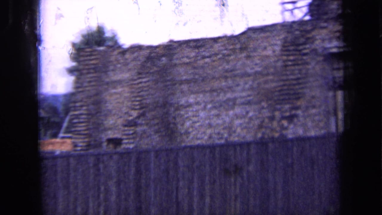 1963 Russia home-movie frame with rough brickwork, a high wooden fence, partial scaffolding, and trees beyond the wall.