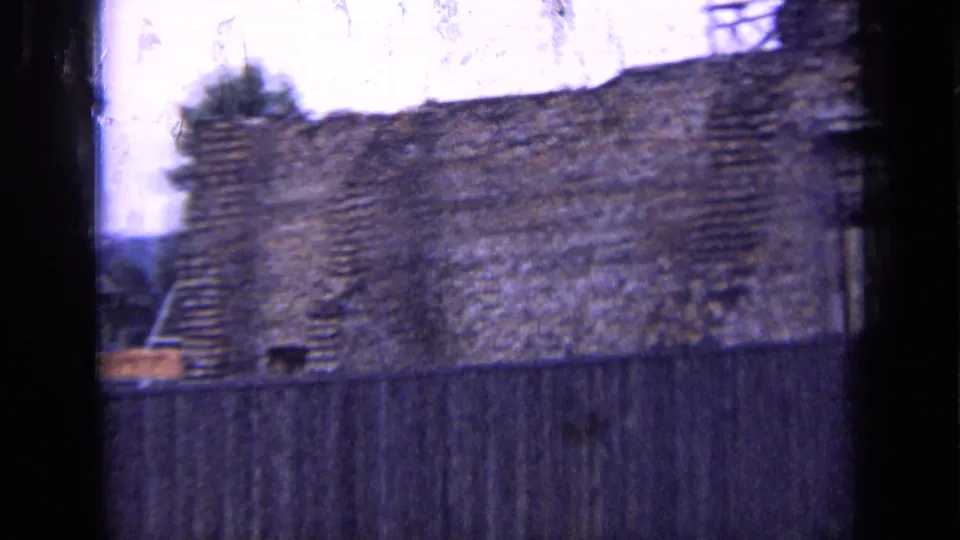 1963 Russia home-movie frame with rough brickwork, a high wooden fence, partial scaffolding, and trees beyond the wall.