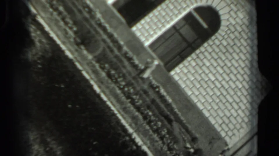 1963 Russian 8mm frame with a tilted view of a brick exterior, garden edge, and shadowed ground.