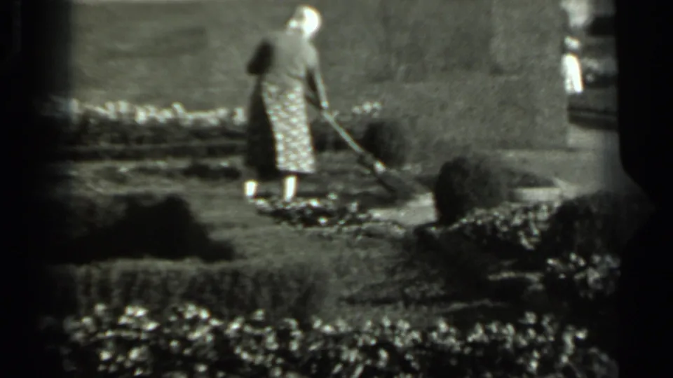 1963 Russian home-movie frame of a woman in a white hat and patterned skirt sweeping a sunny garden path.
