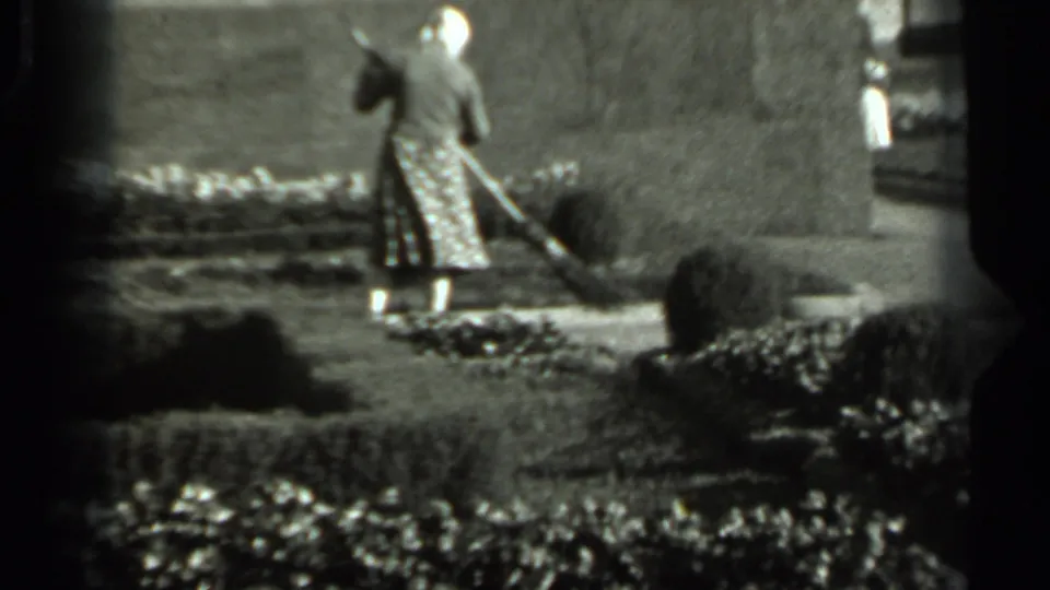 In 1963 Russia home-movie footage, a woman in a head covering and patterned dress sweeps near hedges and a garden wall.