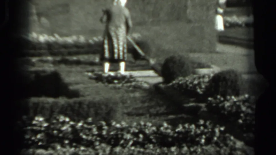 The woman pauses or turns with a broom on a garden path, with manicured hedges and a wall visible in the 1963 Russia footage.
