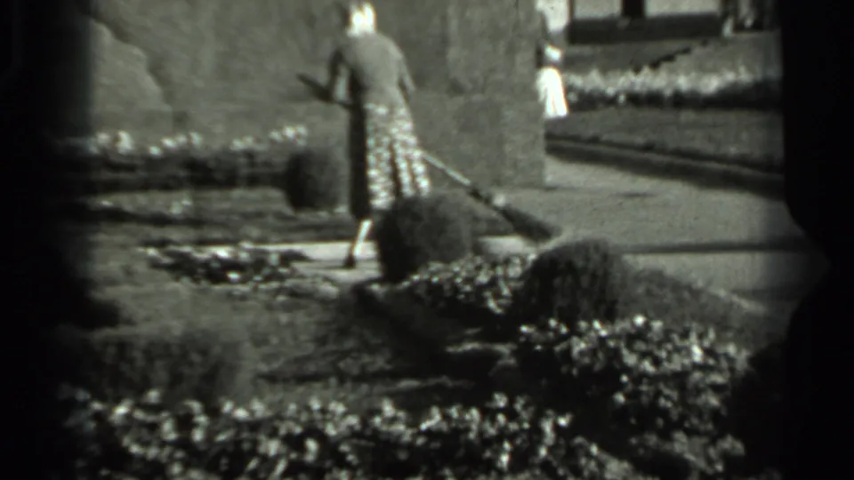 A woman in a patterned dress sweeps the path while a second figure is briefly visible behind garden walls or plantings.