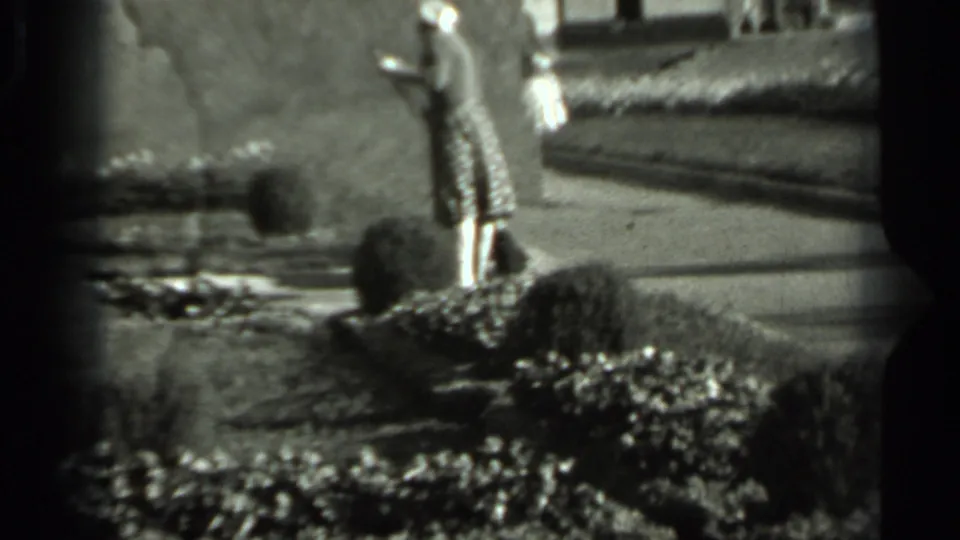 The woman stands with a broom near clipped hedges and a paved or gravel path, recorded in soft 8mm black-and-white footage.