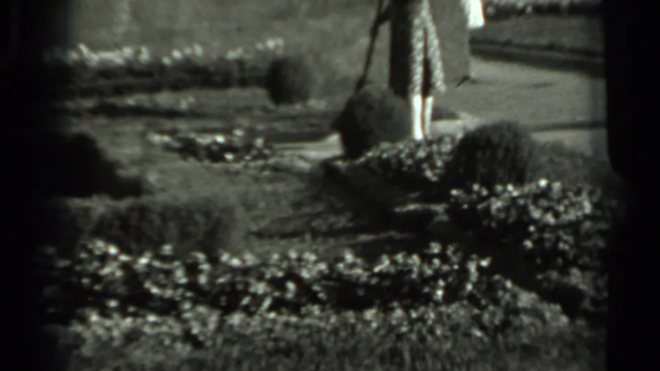 1963 Russia home-movie frame of a woman in a floral dress sweeping near clipped shrubs and a paved garden path.