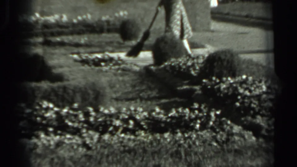 A woman in a patterned dress continues sweeping around manicured garden beds in black-and-white 8mm footage from Russia.