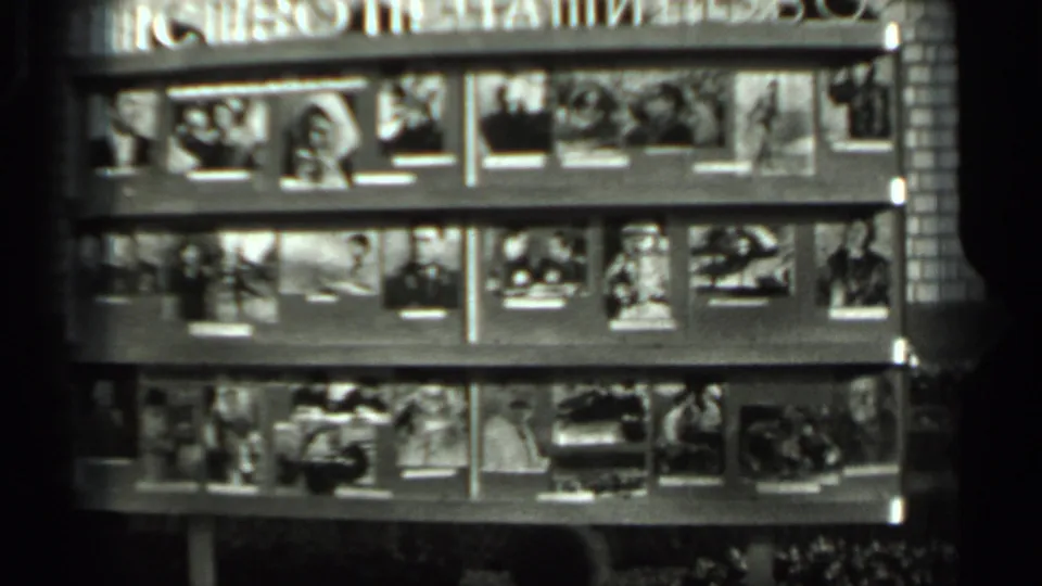 Black-and-white 8mm frame showing a grid-like wall display of personal photographs, with individual faces visible in some prints.