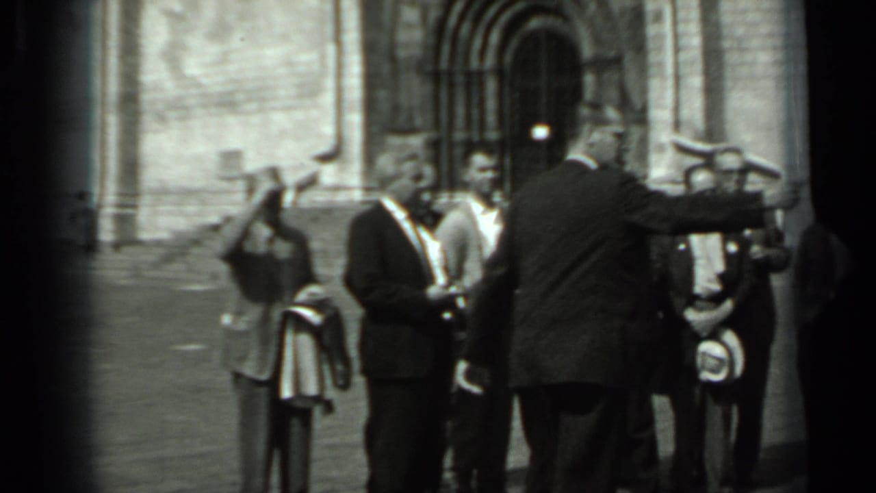 In 1963 Russia, a group of men in suits appears to pose outside a large stone building as one man gestures toward them.