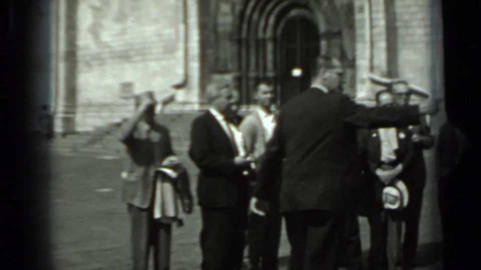 In 1963 Russia, a group of men in suits appears to pose outside a large stone building as one man gestures toward them.