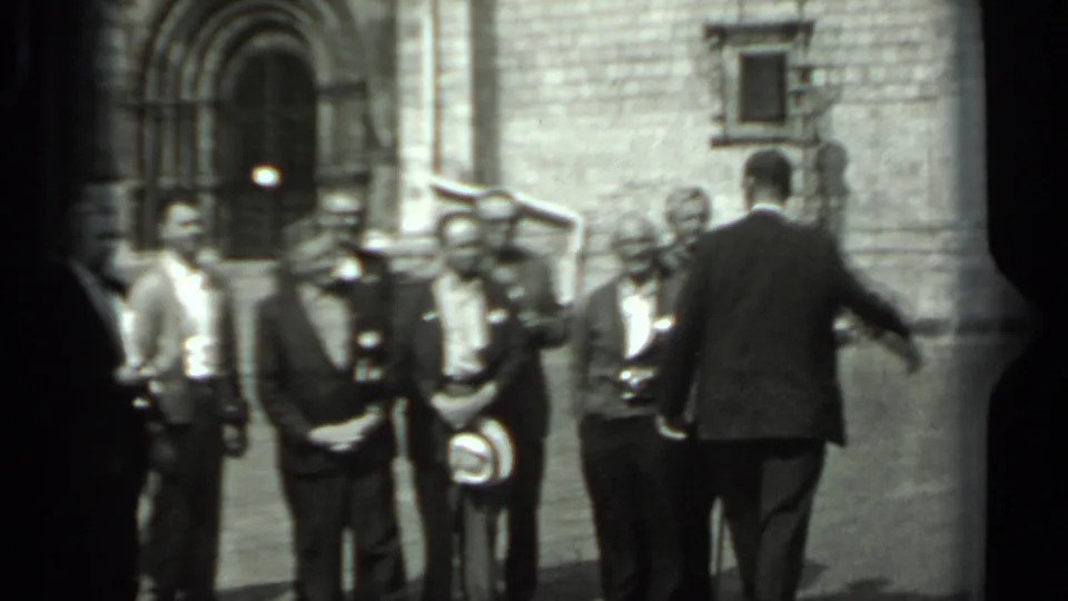 A man walks or turns across the foreground as the suited group holds its pose near the arched stone entrance.