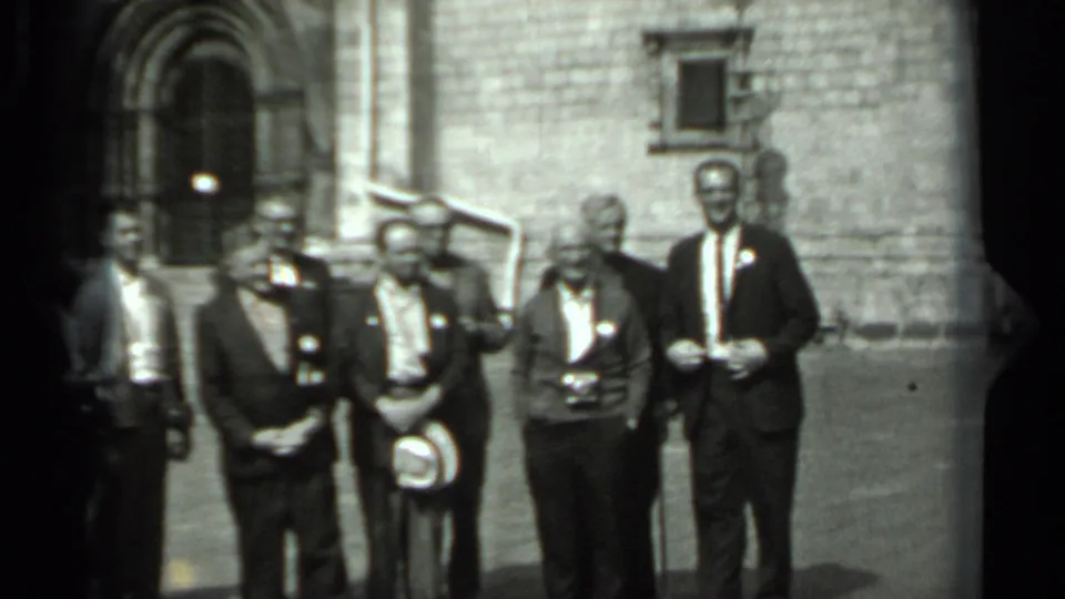 The group stands more evenly arranged for the camera, with suits, ties, held hats, and the stone courtyard visible behind them.