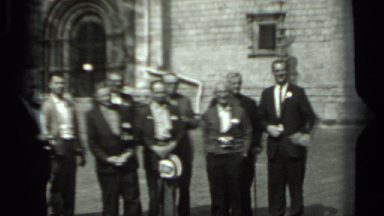 In 1963 Russia footage, men in suits and badges stand before a historic stone facade with an arched entrance.