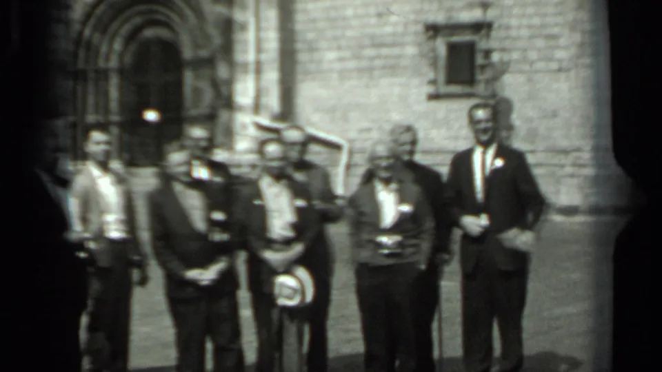 In 1963 Russia footage, men in suits and badges stand before a historic stone facade with an arched entrance.