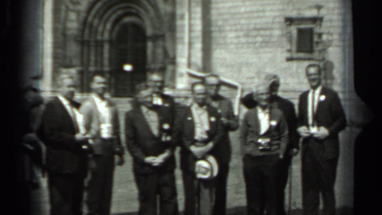 A line of men stands near a stone building entrance in daylight.