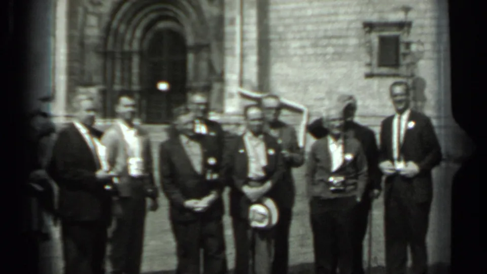 Men, some wearing badges, stand in line outside a Russian stone building during the 1963 home-movie scene.