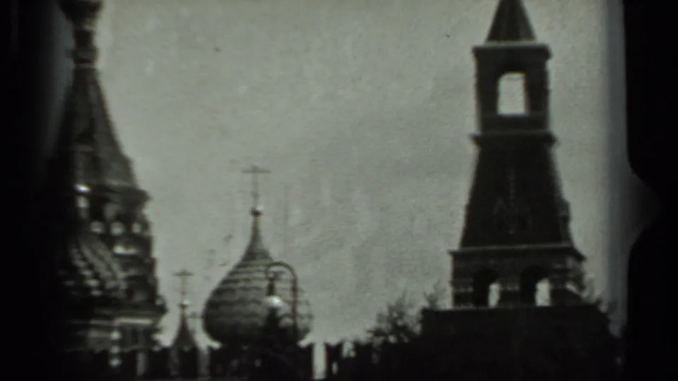1963 Russia 8mm frame with a bell tower, onion domes, crosses, and dark foreground trees in a visitor-shot view.
