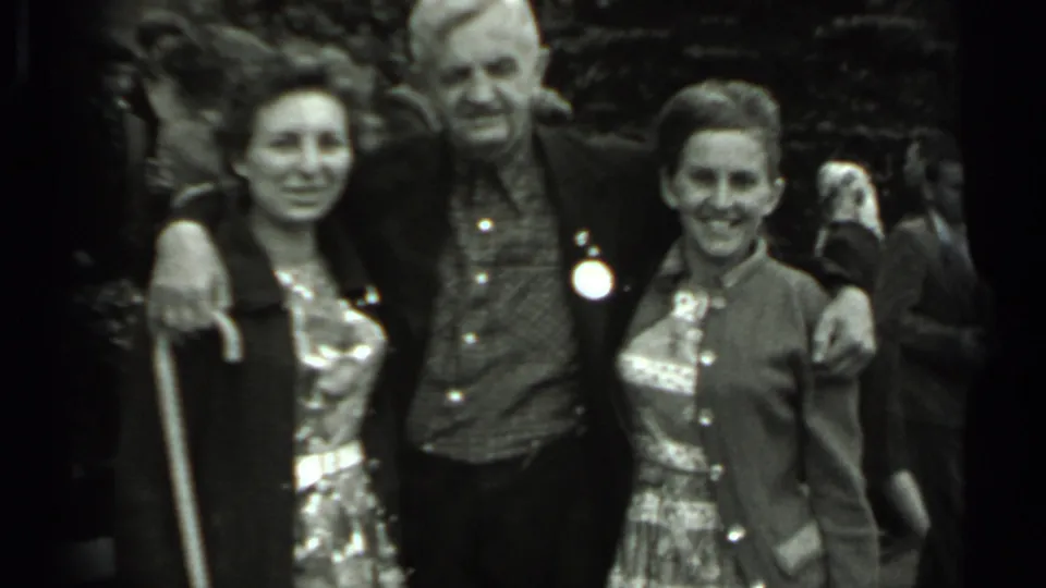 In 1963 Russia home-movie footage, an older man stands with his arms around two women as they smile in a crowded outdoor setting.