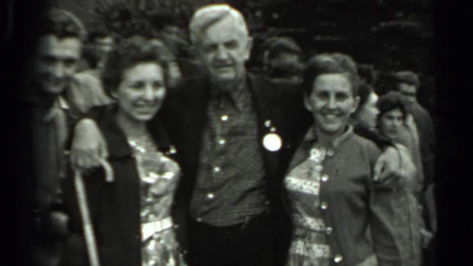 The handheld 8mm frame shows a group portrait in Russia, with an older man between two women and another man partly entering the shot.