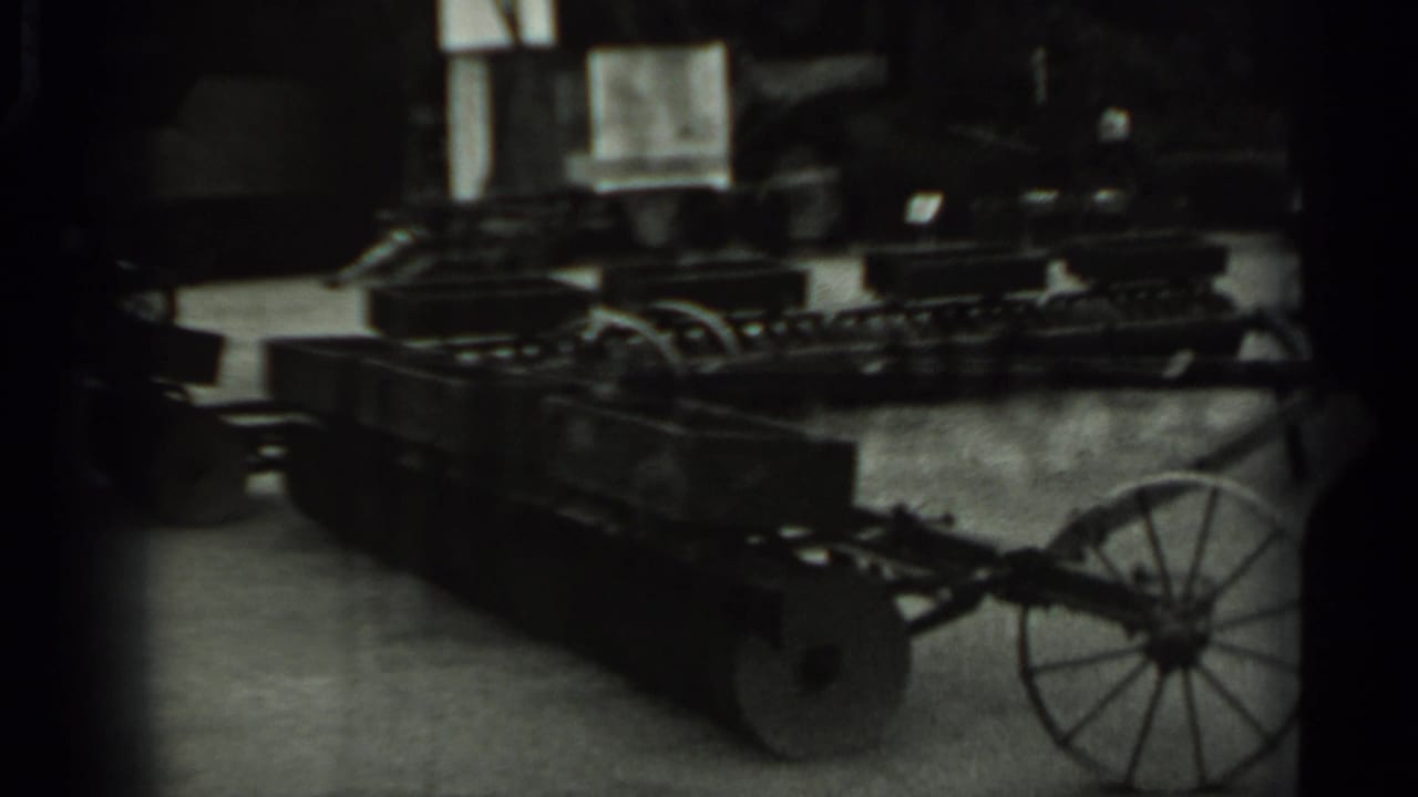 Long wheeled metal farm cart indoors beside stacked equipment and windows.
