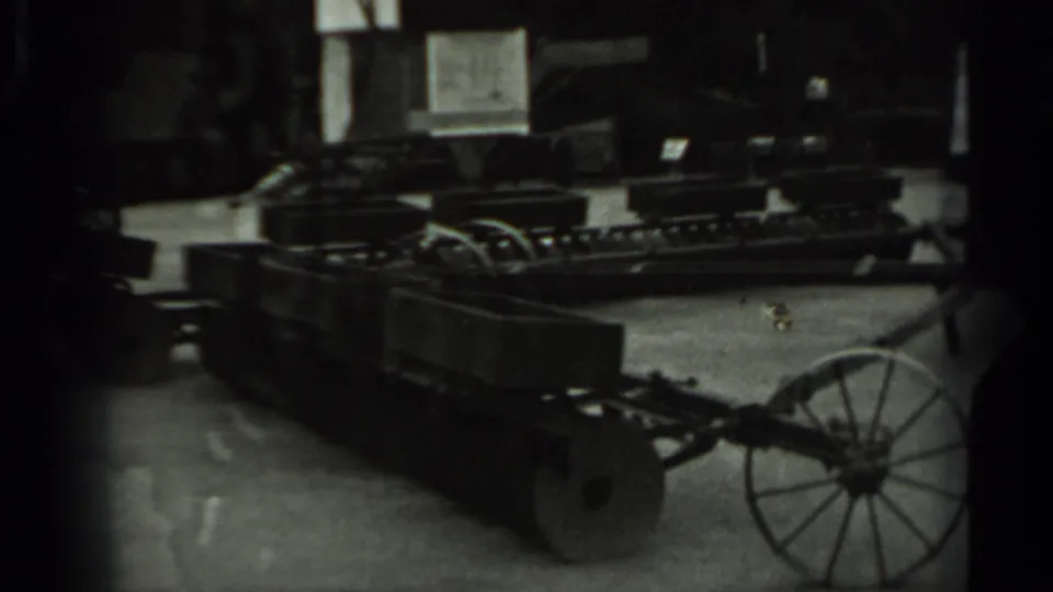 Black-and-white 8mm view of a wheeled agricultural feeder or transport cart stored inside, with equipment and window light behind it.