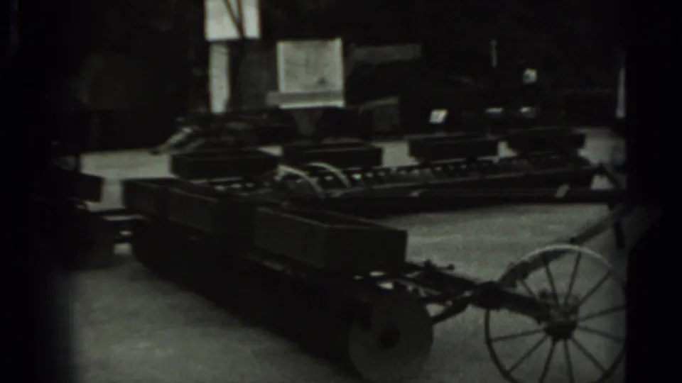 Monochrome 8mm frame showing a wheeled machine or cart-like apparatus, with a large spoked wheel and shadowed mechanical parts.