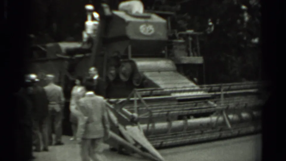1963 Russia frame showing adults in period clothing gathered near a large agricultural machine, likely a combine harvester, with trees behind it.