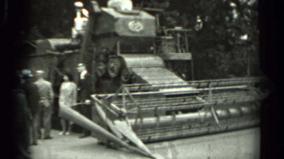 1963 Russia 8mm frame with suited adults viewing a large agricultural machine, railings, trees, and a visible emblem or number on the equipment.