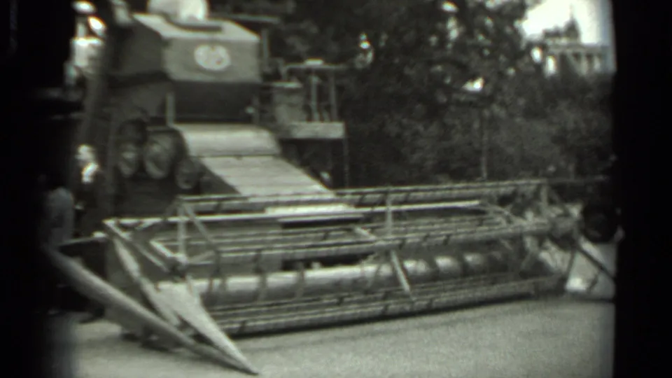 Black-and-white 8mm frame of a combine harvester display in Russia, with visitors partly visible near the front machinery.