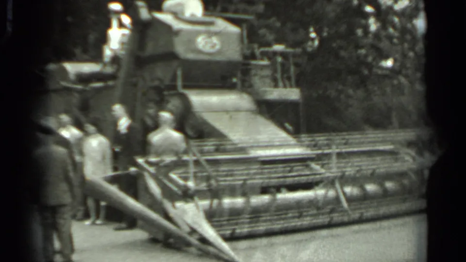 Black-and-white 8mm frame from Russia showing a small group beside a displayed combine harvester and its front mechanism.