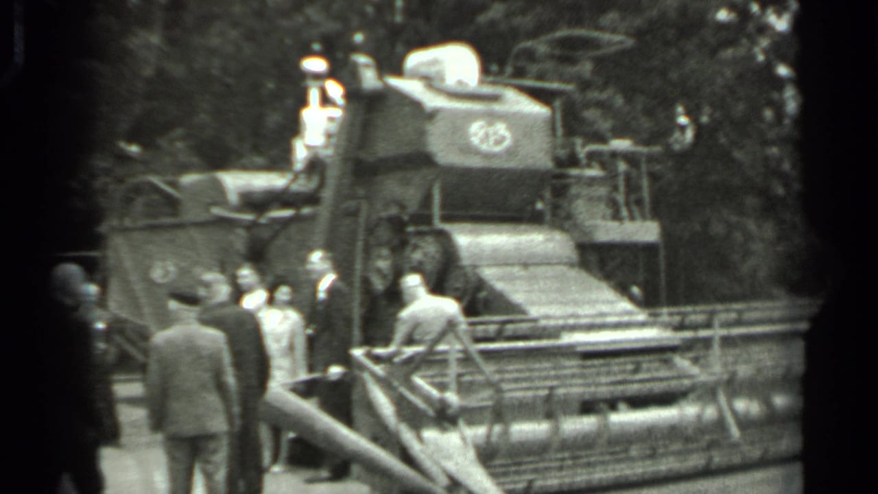 Group of men stand near the side of a large harvester with trees behind it.